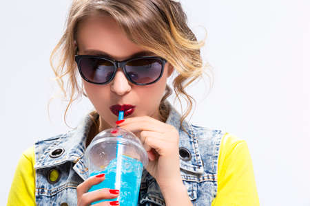 Happy Youth Lifestyle Concepts. Closeup of Upbeat Caucasian Blond Girl Drinking Blue Cocktail Through The Straw. Wearing Denim Vest and Sunglasses.Horizontal Imageの写真素材