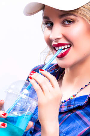 Lifestyle Youth Ideas. Portrait of Sexy Smiling Caucasian Blond Woman in Checked Shirt Drinking Blue Juice Through Straw. Wearing Sun Visor. Vertical Image Compositionの写真素材