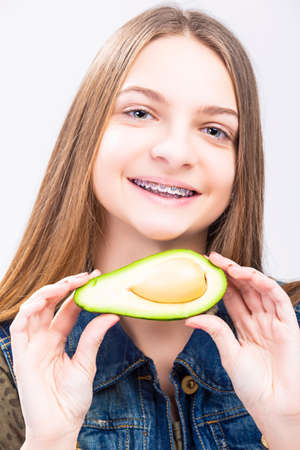 Portrait of Smiling Caucasian Female Teenager With Teeth Braces. Posing with Long Green Avocado Fruit. Vertical Shotの写真素材