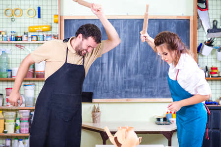 Two Caucasian Ceramists Squarrel and Crashing Clay Bowl on Table in Front with rolling Pins. Negative and Furious Facial Expression.Horizontal Shotの写真素材