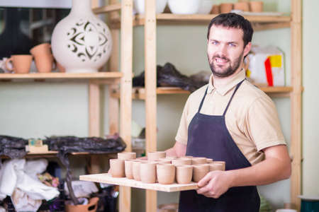 Portrait of Professional Male Ceramist Holding Tray with Clay Handmade Cups. Posing in Protective...