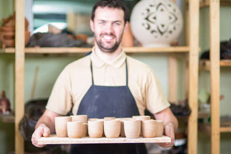 Portrait of Professional Male Ceramist Holding Wooden Tray with Clay Handmade Cups. Posing in...