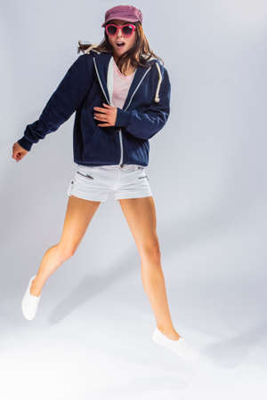 Portrait of Happy Jumping Caucasian Brunette Woman in Classic Hoodie and Rose Cap with Sunglasses. Posing Against White Background. Vertical imageの写真素材