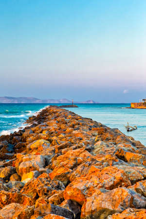 Long Stony Spit Going Far to Sea in Heraklion City on Crete, Greece. Roaring Waves Batter Against The Rocks During Golden Hour. Vertical Imageの写真素材