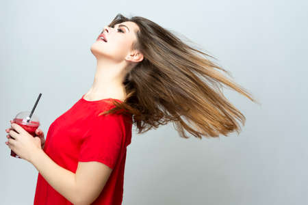 Positive Caucasian Female Dancing with Cup of Red Smoothie Juice Mix. Posing in Red Dress Over White. Horizontal Imageの写真素材