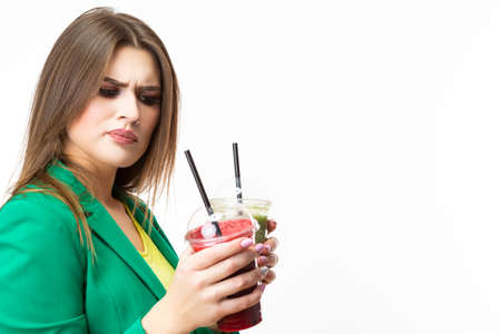 Healthy Food Eating. Choosy Thinking Woman With Both Green and Red Detox Vegetable Smoothie. Posing in Green Jacket Over White.Unpleased Facial Expression. Horizontal Shotの写真素材