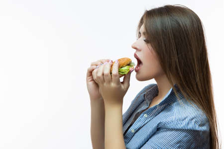 Unhealthy Eating Concepts. Caucasian Woman Eating Burger. Profile Face View. Posing in Striped Shirt Indoors in Studio. Horizontal Image Compositionの写真素材