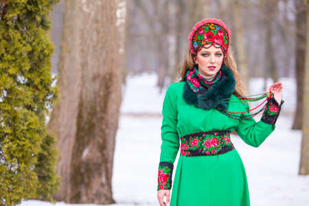 Caucasian Blond Girl in Fashionable Green Dress and Kokoshnik with Flowery Pattern and Beads. Posing in Winter Snowy Park Outdoors. Horizontal Image Compositionの写真素材