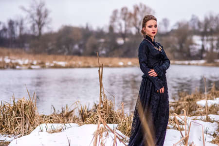 Young Caucasian Brunette Female in Art Dress with Necklace and Crown Posing Against River Outdoors....