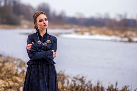 Gorgeous Caucasian Brunette Female in Art Dress with Necklace and Crown Posing Against River...