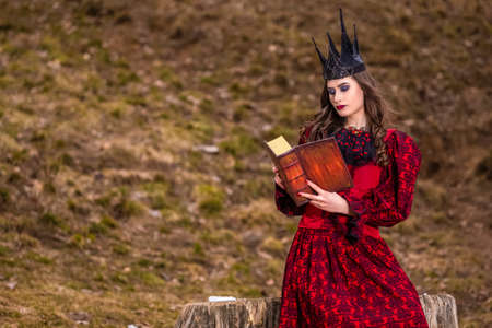 Art Photography. Mysterious Fairy Medieval Queen in Red Dress and Spiky Black Crown Posing With Ancient Book in Forest in Early Spring.Horizontal Imageの写真素材