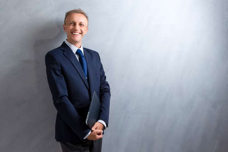 Portarit of Smiling Caucasian Man in Blue Suit Posing with Laptop Against Grunge Wall. Horizontal Image Compositionの写真素材