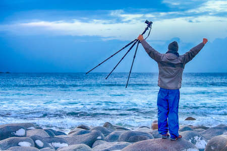 Man With Tripod and Camera Standing on Rocky Ocean Shore with Lifted Hands.Horizontal Image...