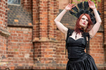 Gothic Woman in Black Dress and Feather Crown.Against Brick Wall Outdoors. Posing with Lifted Hands....