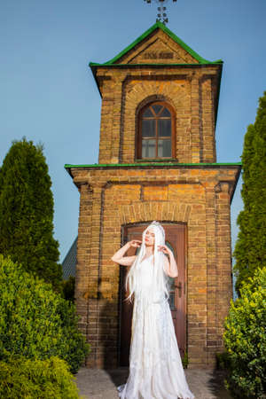 Young Caucasian Bride with Long White Hair Posing Against Old Brick Church Outdoors. Vertical Imageの写真素材