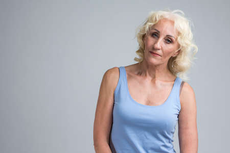 Natural Portrait of Tranquil Mature Blond Woman in Blue Tank Top Posing Against White Background in Studio. Horizontal Imageの写真素材
