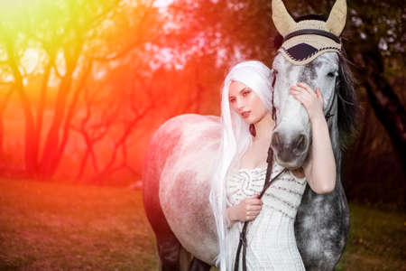 Beautiful Caucasian Blond Female with long Hair Standing with Grey Horse Against Bright Light on The...