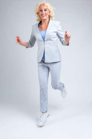 Happy Mature Blond Woman in Fashionable Suit Jumping and Moving Against White Background in Studio.Vertical Image compositionの写真素材