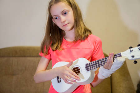 Portrait of Caucasian Girl  With Injured Hand In Plaster. Posing with Small Guitar Indoors....