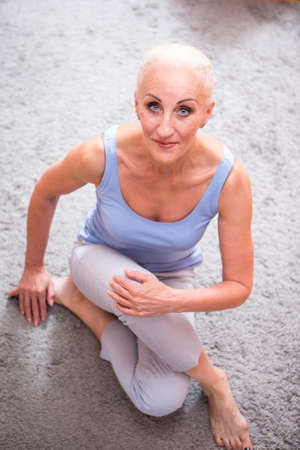Mature Adults Concepts. Portrait of Positive Caucasian Blond Senior Woman Posing in Casual Clotthing. Sitting on Floor with folded Legs. Vertical Compositionの写真素材