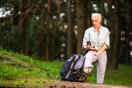 Portrait of Relaxing Senior Woman Posing with Smartphone and Tourist Backpack in Forest Outdoors. Horizontal Imageの写真素材