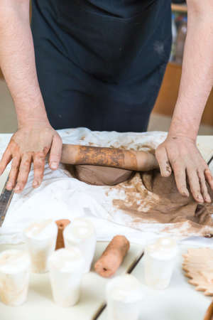 Pottering Concepts. Closeup of Hands of Male Worker Rolling a Piece of Wet Clay on Table Before...