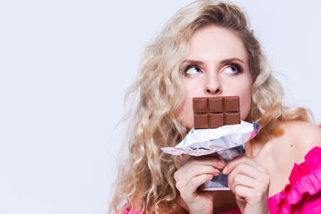 Portrait of Cute Blond Caucasian Female Eating Chocolate Bar. Posing on White. Horizontal Imageの写真素材
