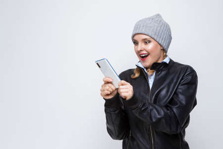 Youth Lifestyles.Excited Caucasian Blond Femalr in Warm Hat and Leather Jacket Posing With Cellphone...