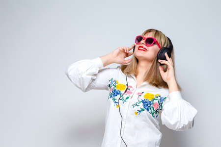 Youth Lifestyle Concepts. Portrait of Happy Caucasian Blond Girl In Shorts and Red Glasses. Lislting to Music in Headphones on White. Horizontal Image Compositionの写真素材