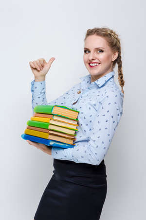 Education Concepts and Ideas. Caucasian Blond Female Posing With Heap of Books in Colorful Covers With Pleased Facial Expression. Showing Thumbs Up Sign Against White. Vertical Imageの写真素材