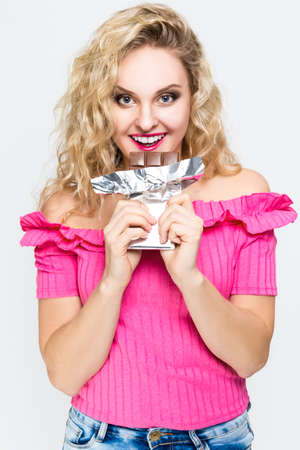 Food Concepts. Portrait of Cute and Charming Caucasian Blond Female Eating Chocolate Bar. Posing on White. Vertical Imageの写真素材