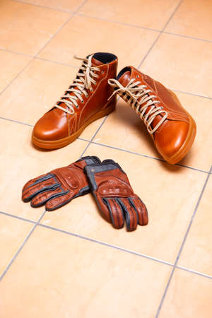 Motorcycling Relative Topics. Closeup of Protective Motorcyclist Leather Tan Sneakers Placed Indoors Along With Leather Gloves On Pale Tiles Floor. Vertical Image compositionの写真素材