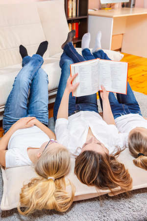 Family Ideas. Portrait of Caucasian Mother Reading a Book Together with Her Female Twins Laying on Floor Indoors. Vertical Compositionの写真素材