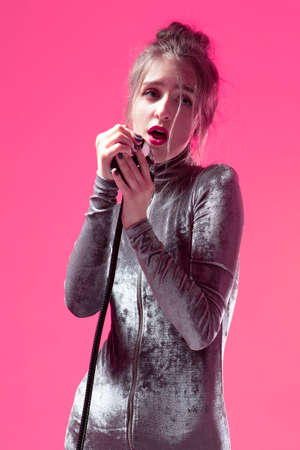 Funny Singing Caucasian Girl In Gray Reflective Dress Posing With Microphone Indoors On Pink Background. Vertical Imageの写真素材