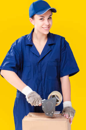 Shipment Ideas. Smiling Caucasian Female Courier in Coverall Packaging Box with Tape Device. Against Yellow Background. Vertical Imageの写真素材
