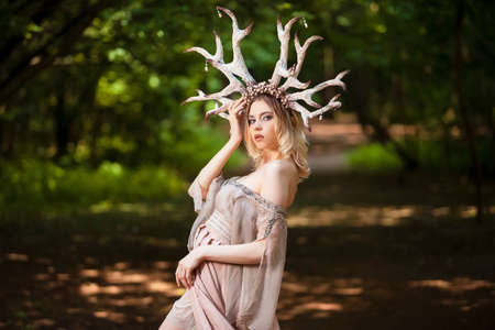 Beautiful Girl Posing With Artistic Deer Horns  In Summer Forest Near High Tree. Wearing Light Dress for Forest Nymph Concept. Art Photography. Vertical Imageの写真素材