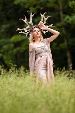 Beautiful Caucasian Girl With Artistic Deer Horns In Front of Tree in Forest. Posing in Light Dress...