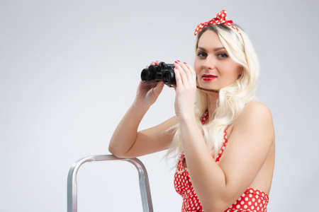 Blond Girl Pin-up Style Posing With Binoculars Near Stepladder. Looking Upwards.Posing  Against White. Horizontal Image Compositionの写真素材
