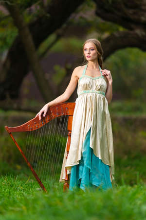 Natural Portrait of Sensual Female Harpist Woman in Light Dress Posing With Harp  In Park Outdoor....