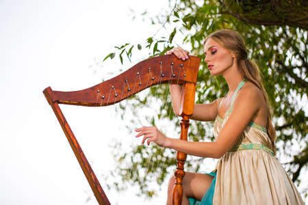 Natural Portrait of Cute Caucasian Blond Girl in Long Dress Playing the Harp on tree Branch on Park...