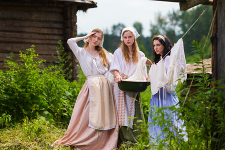 Group of Three Beautiful Caucasian Girlfriends Standing Together in Traditional Rural Costumes With Basin of Linens at Countryside Outdoor. Horizontal Imageの写真素材