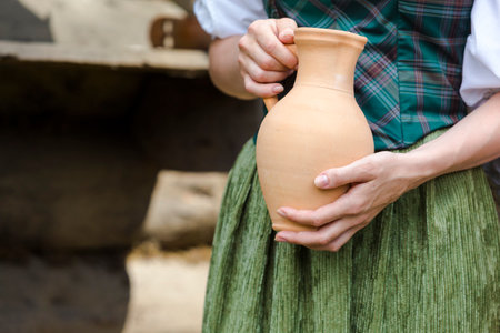 Closeup of Hands of Caucasian Girl Holding Clay Jar in Front of Decorated Dress Outdoor.の写真素材