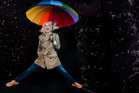 Full Length Portrait of Smiling Caucasian Blond Female Jumping with Colorful Umbrella Under The Multiple Water Dropletsの写真素材