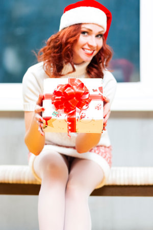 Holidays Concepts. Portrait Lovely  Cute Caucasian Santa Helper Girl Posing in Santa Hat Offering Christmas Gift Box. Vertical Composition. Focus on Box.の写真素材