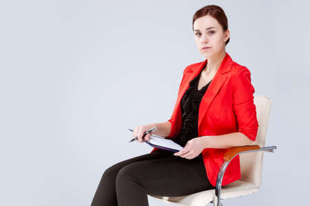 Women in Business Ideas. Portrait of Caucasian Brunette Enterpreneur in Red Blazer Posing in Chair with Notebook. Horizontal Image Compositionの写真素材