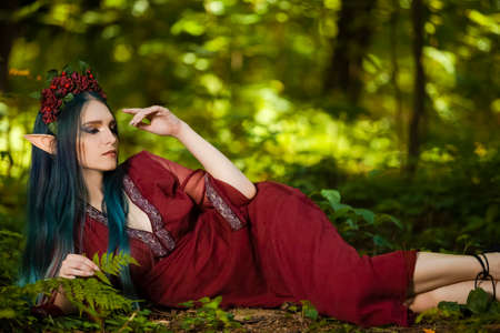 Portrait of Cute Caucasian Girl in Flowery Garland and Artistic Elf Ears Posing on Ground in Summer Forest. Horizontal Image Compositionの写真素材