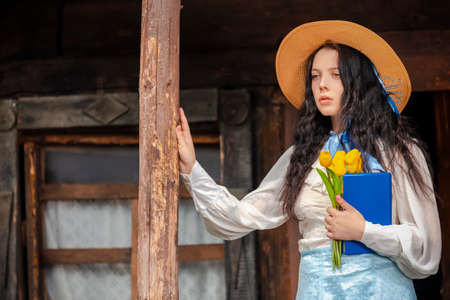 Portrait of Dreaming Caucasian Brunette Girl in Old-fashioned Dress With Blue Book and Yellow Flowers In Front of Old Wooden House. Horizontal Image Compositionの写真素材