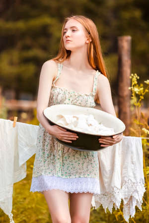 Redhaired Caucasian Girl Posing With Basin of Laundry Against Hanged Linen on  Rope Behind In Countryside Environment.Vertical Imageの写真素材
