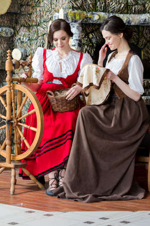 Portrait of Two Young Girls Posing With Old Wooden Spinning Wheel in Retro Dress In Rural Environment.Vertical Imageの写真素材