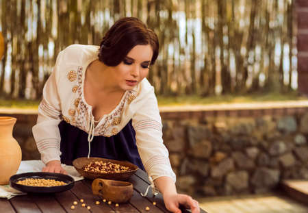 Portrait of Sexy Caucasian Female in Rural Decorated Dress Posing with Wooden Plates Filled with Frigole Beans In Rural Environment. Horizontal Imageの写真素材